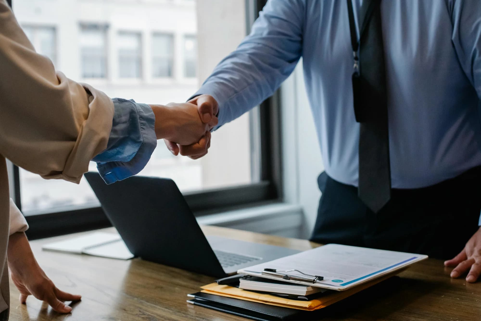 a man in a blue shirt shaking hands with a client over a table with a laptop on it